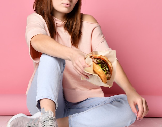 Stylowa i młoda kobieta imbirowa trzyma kanapkę z burgerem na różowym tle