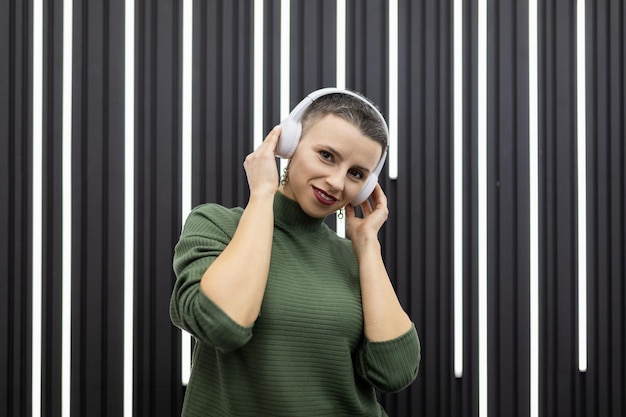 Stylowa kaukaska kobieta na tle szarej ściany słucha muzyki w słuchawkach z uśmiechem