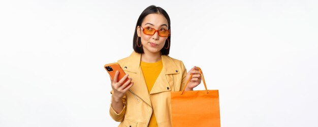 Stylowa klientka azjatyckiej dziewczyny nosi okulary przeciwsłoneczne, trzymając torbę na zakupy i smartfona, idąc po zniżki w sklepach stojących na białym tle