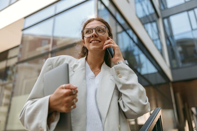Stylowa kobieta biznesowa stojąca z laptopem na tle budynku biurowego i rozmawiająca przez telefon