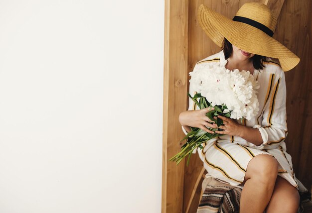 Zdjęcie stylowa kobieta boho w kapeluszu trzymająca bukiet białych piwonii na rustykalnym drewnianym tle miejsce na tekst hipster dziewczyna w sukience pozuje z piwoniami szczęśliwy dzień matki międzynarodowe dni kobiet