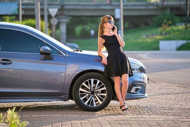 Stylowa kobieta kierowca stojący w pobliżu swojego pojazdu, rozmawiający przez telefon komórkowy na ulicy miasta w lecie