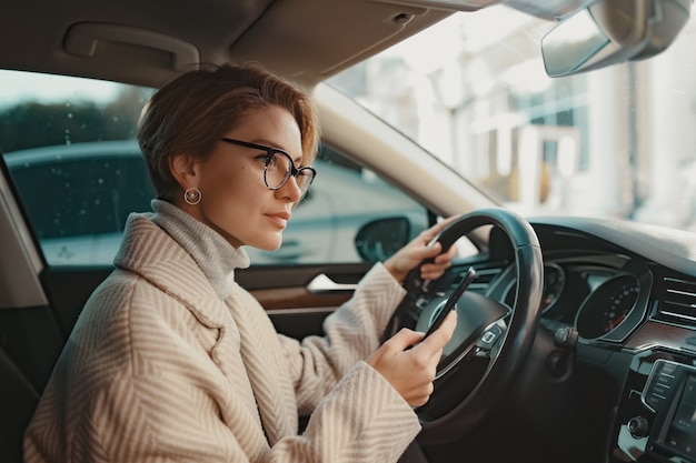 stylowa kobieta siedząca w samochodzie ubrana w zimowy płaszcz i okulary za pomocą smartfona