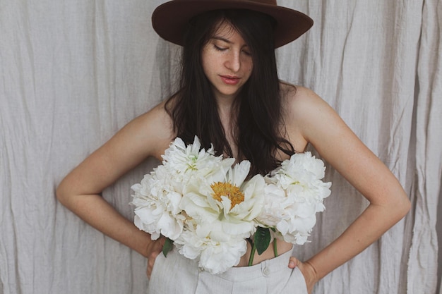 Stylowa kobieta w kapeluszu pozuje z białymi kwiatami piwonii wyrastającymi ze spodni Kwiatowy zapach Self care