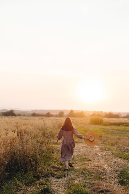 Stylowa kobieta w słomkowym kapeluszu spacerująca po polu owsa w świetle zachodzącego słońca widok z tyłu Atmosferyczna spokojna chwila Młoda kobieta w lnianej sukience relaksująca się wieczorem na wsi wiejskie powolne życie