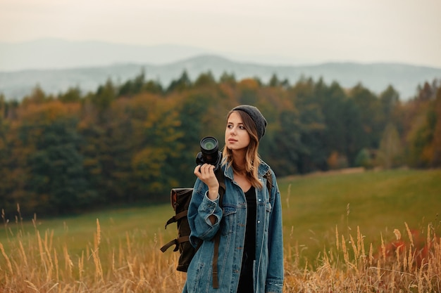 Stylowa kobieta z aparatem fotograficznym i plecakiem na wsi z górami