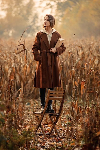 Stylowa kobieta z książkowym pobytem na krześle na kukurydzanym polu w jesień czasu sezonie