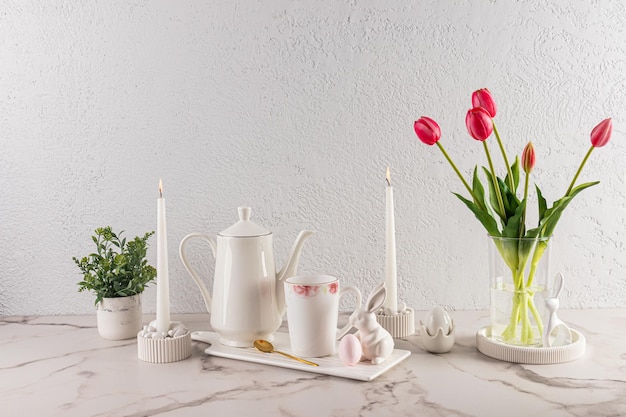 Stylowa martwa natura na święta wielkanocne Biały dzbanek do kawy na ceramicznej białej tacy z kubkiem figurki zajączka wielkanocnego wazon z kwiatami i świecami
