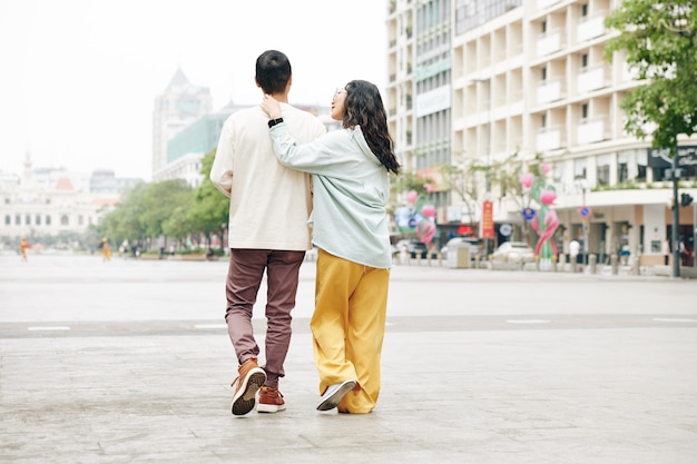 Stylowa młoda Chinka dotyka szyi chłopaka, gdy idą po placu miejskim, widok z tyłu