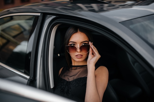 Stylowa młoda dziewczyna siedzi w samochodzie klasy biznes w czarnej sukience. Biznesowa moda i styl