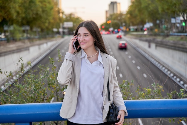 Stylowa młoda kobieta biznesu używa smartfona stojącego przy drodze na ulicy miasta o zachodzie słońca