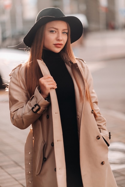 Stylowa młoda kobieta w beżowym płaszczu w czarnym kapeluszu na ulicy miasta. Moda uliczna dla kobiet. Jesienna odzież. Miejski styl.