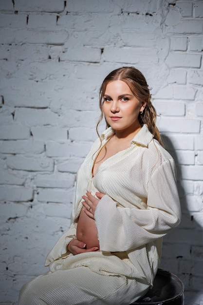 Stylowa młoda kobieta w ciąży w białym garniturze siedzi pod białą ceglaną ścianą i delikatnie dotyka brzucha Oczekiwanie na narodziny Opieka i macierzyństwo Miłość i czułość