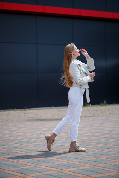 Stylowa młoda kobieta z długimi blond włosami o europejskim wyglądzie z uśmiechem na twarzy. Dziewczyna w białej kurtce i białych dżinsach ciepły letni słoneczny dzień na tle szarego budynku