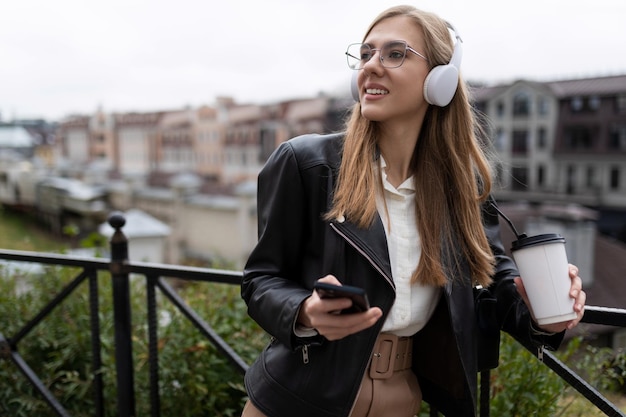 Stylowa młoda studentka w słuchawkach trzymająca telefon i filiżankę kawy w dłoniach na zewnątrz