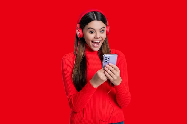 Stylowa nastolatka słucha muzyki w słuchawkach wybiera piosenkę na telefonie izolowana na czerwonym