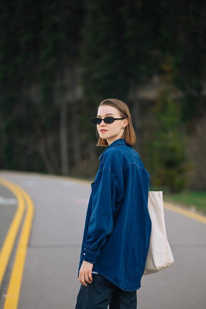 stylowa pani w niebieskiej koszuli z torbą w rękach i okularami przeciwsłonecznymi spaceruje górską drogą