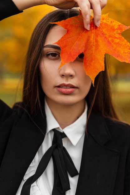 Stylowa piękna elegancka kobieta w modnych czarnych ubraniach biznesowych z płaszczem i krawatem trzyma kolorowy czerwony jesienny liść klonu w pobliżu twarzy w jesiennym parku