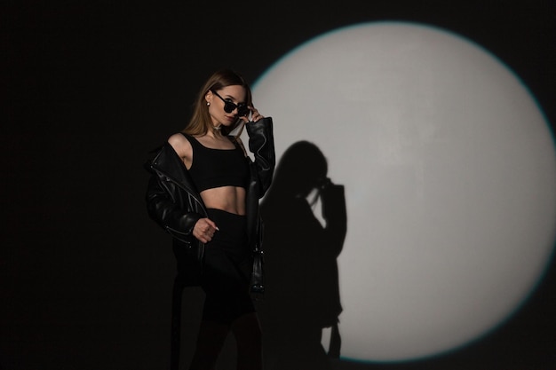 Stylowa piękna hipster dziewczyna w modnym czarnym rockowym stroju zakłada okulary przeciwsłoneczne na ciemnym tle w studio na imprezie