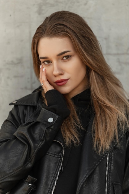 Stylowa piękna młoda dziewczyna modelka hipster w modnych ubraniach z czarną skórzaną kurtką i czarną bluzą z kapturem na ulicy w pobliżu szarej betonowej ściany vintage