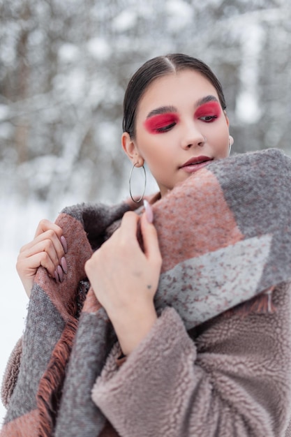 Stylowa piękna młoda dziewczyna z różowymi cieniami do powiek i kolorowym makijażem w modnych zimowych ubraniach z futrem i szalikiem wychodzi na zewnątrz ze śniegiem