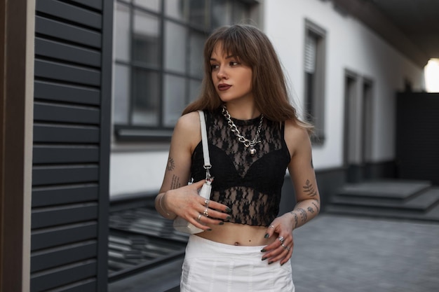 Stylowa piękna młoda kobieta hipster z tatuażem na ramionach w modnym letnim seksownym stroju z torebką chodzi w pobliżu budynku na ulicy