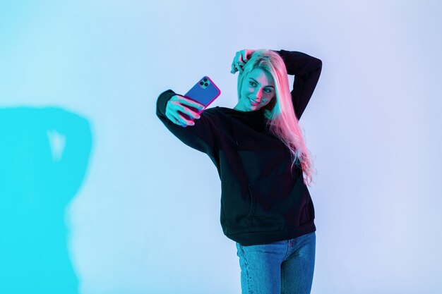 Stylowa piękna młoda kobieta w modnej czarnej bluzie z dżinsami robi selfie na telefonie komórkowym w studio na wielokolorowym neonowym różowym tle. Kreatywny obraz