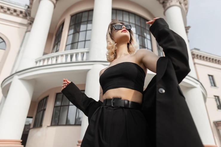 Zdjęcie stylowa piękna młoda kobieta w modnych czarnych eleganckich ubraniach z górną marynarką i spodniami, spacerując i ciesząc się miastem w pobliżu zabytkowego budynku