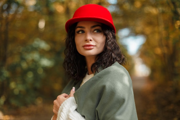 Stylowa piękna młoda ładna kobieta z kręconym czerwonym kapeluszem i modnym białym swetrem z dzianiny z zielonym szalikiem spaceruje po jesiennym parku