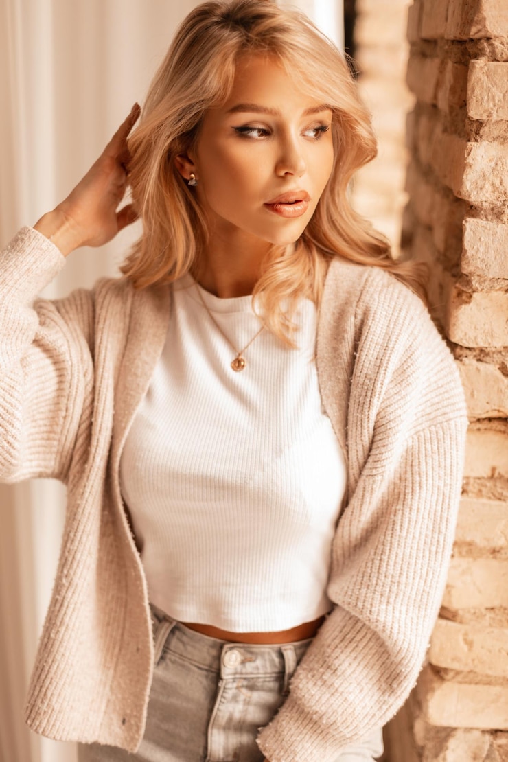 Zdjęcie stylowa słodka kobieta z blond fryzurą w modzie dzianinowy sweter biała koszulka i dżinsy stoi w pobliżu ceglanego muru w pomieszczeniu