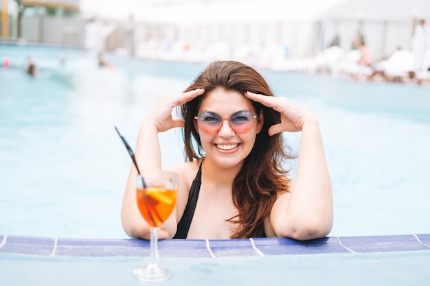 Zdjęcie stylowa szczęśliwa młoda kobieta w rozmiarze plus size w czarnym stroju kąpielowym i okularach przeciwsłonecznych, ciesząca się życiem ze szklanką koktajlu w hotelowym basenie na wakacjach