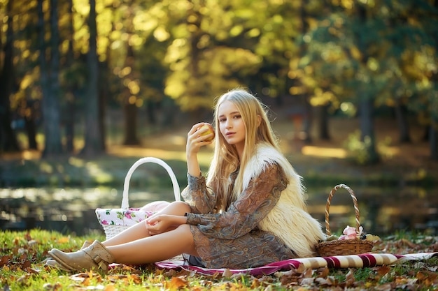 Stylowa zmysłowa przemyślana piękna młoda blond dziewczyna z długimi włosami w modnej wiosennej sukience siedzącej na piknikowej kratę z koszem owoców w parku na świeżym powietrzu na naturalnym tle poziomy obraz
