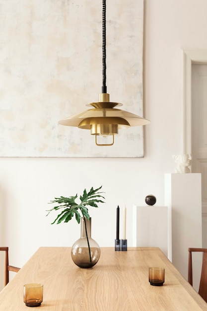 Stylowe i nowoczesne wnętrze jadalni z designerskim stołem, krzesłami, złotą lampą wiszącą, abstrakcyjnymi obrazami i eleganckimi dodatkami. Tropikalne liście w wazonie. Eklektyczny wystrój domu.