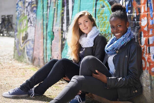 Zdjęcie stylowe kobiety siedzące na zewnątrz przy ścianie w kampusie i uśmiechnięte młode przyjaciółki szczęśliwe, różnorodne studentki, cieszące się więzią lub dobrym czasem, modą lub modą i bawiące się w dzień wolny