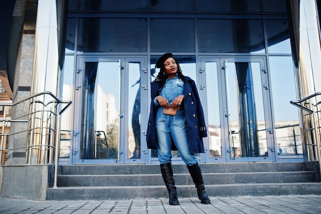 Stylowe modne afroamerykanki w dżinsach i czarnym berecie z niebieskim płaszczem na tle nowoczesnego budynku