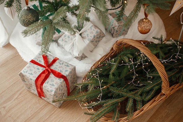 Stylowo owinięte prezenty bożonarodzeniowe i rustyczny koszyk z gałęziami sosny pod świątecznie ozdobioną choinką w skandynawskim pokoju Wesołych Świąt i Wesołych Święt