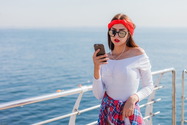 Stylowo ubrana młoda kobieta słucha muzyki w słuchawkach i używa smartfona na plaży przed morzem. Moda uliczna