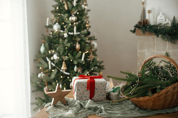 Stylowo zapakowane prezenty bożonarodzeniowe wiejski koszyk z gałęziami sosny i nowoczesnymi dekoracjami na stole w świątecznie ozdobionym skandynawskim pokoju Wesołych Świąt i Wesołych Święt