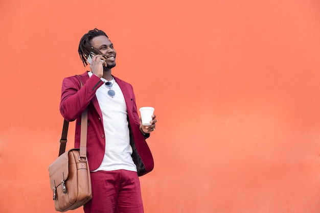 Stylowy afrykański biznesmen ubrany w garnitur z teczką i rozmowami o kawie na smartfonie przed czerwonym tłem, skopiuj miejsce na tekst, pojęcie technologii i komunikacji