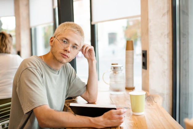 Stylowy blond nastoletni chłopak 18-19 lat nosi okulary do czytania papierowej książki w kawiarni z kawą w filiżance