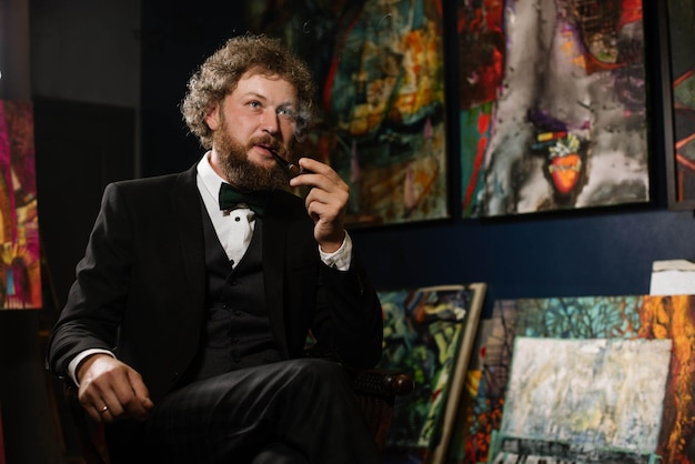 Stylowy brodaty mężczyzna malarz artysta palący fajkę w pobliżu swoich obrazów