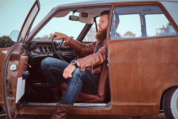 Stylowy brodaty mężczyzna ubrany w brązową skórzaną kurtkę siedzi za kierownicą tuningowanego samochodu retro z otwartymi drzwiami.