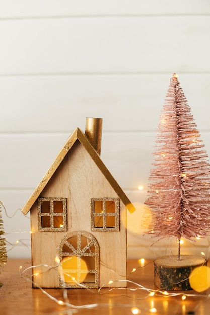 Stylowy dom bożonarodzeniowy i błyszcząca choinka w złotych światłach Nowoczesny świąteczny wystrój w skandynawskim pokoju Miniaturowa wioska z drewnianym domem i zabawkami na drzewie Wesołych Świąt