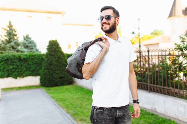 Stylowy hipster z brodą w białej koszulce, z torbą podróżną w rękach spacerującą po mieście
