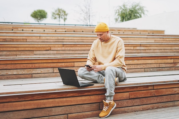 Stylowy mężczyzna hipster w żółtym kapeluszu freelancer pracujący na laptopie w ulicznym parku miejskim