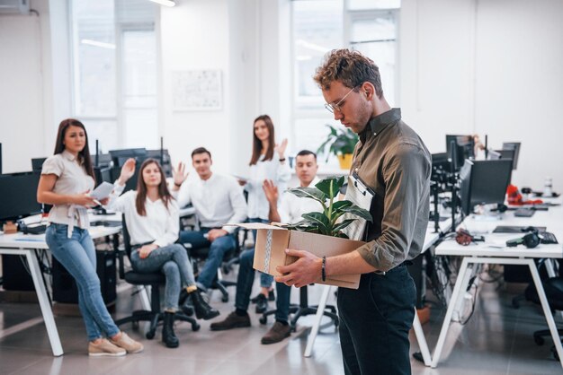 Stylowy mężczyzna w okularach trzymając pudełko z rośliną przed kolegami w biurze.