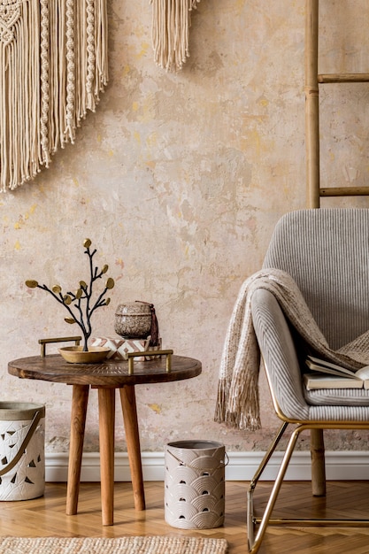Stylowy orientalny salon z designerskim fotelem, piękną makramą, drewnianą drabinką, dekoracją, dywanikiem, stolikiem kawowym, książką i eleganckimi akcesoriami osobistymi w koncepcji wabi sabi.