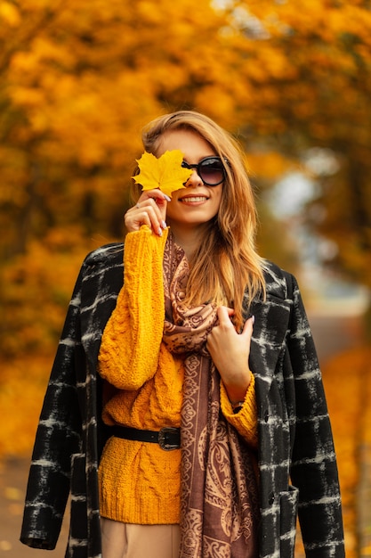 Stylowy portret całkiem szczęśliwej dziewczyny z uśmiechem w okularach przeciwsłonecznych w zabytkowym swetrze z dzianiny, czarnym płaszczu i szaliku zakrywa twarz jesiennym żółtym liściem klonu w niesamowitym parku z liśćmi