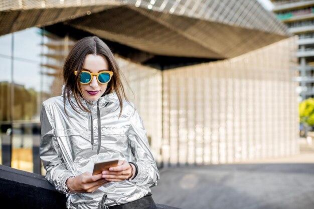 Stylowy portret stylowej kobiety w srebrnej kurtce korzystającej z telefonu na zewnątrz na nowoczesnym tle architektonicznym