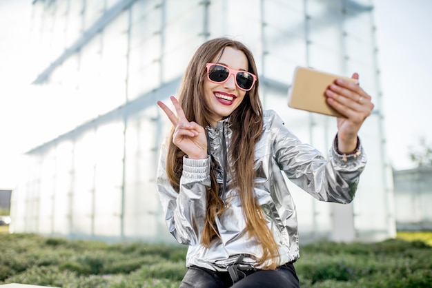 Stylowy portret stylowej kobiety w srebrnej kurtce, która robi selfie w plenerze w nowoczesnym otoczeniu architektonicznym